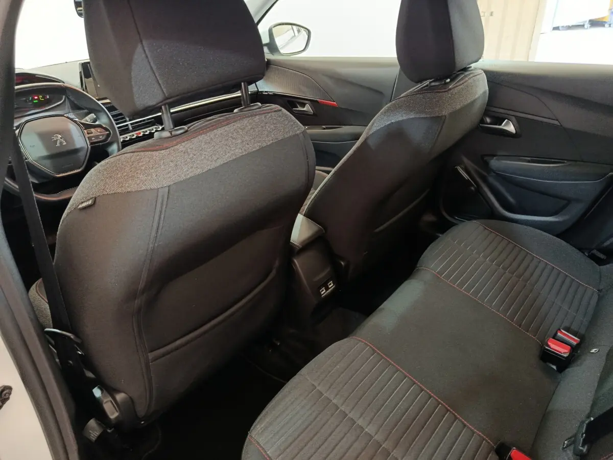 Vue intérieure arrière de la Peugeot 208 électrique blanche, montrant les sièges noirs avec surpiqûres rouges et la console centrale.