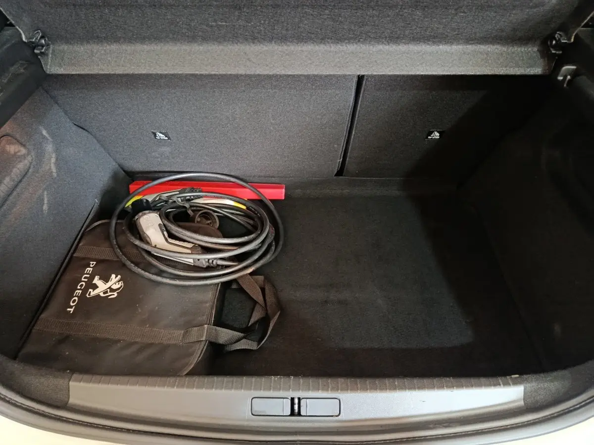 Coffre ouvert d’une Peugeot 208 électrique blanche, avec câble de recharge et sac de rangement Peugeot noir.