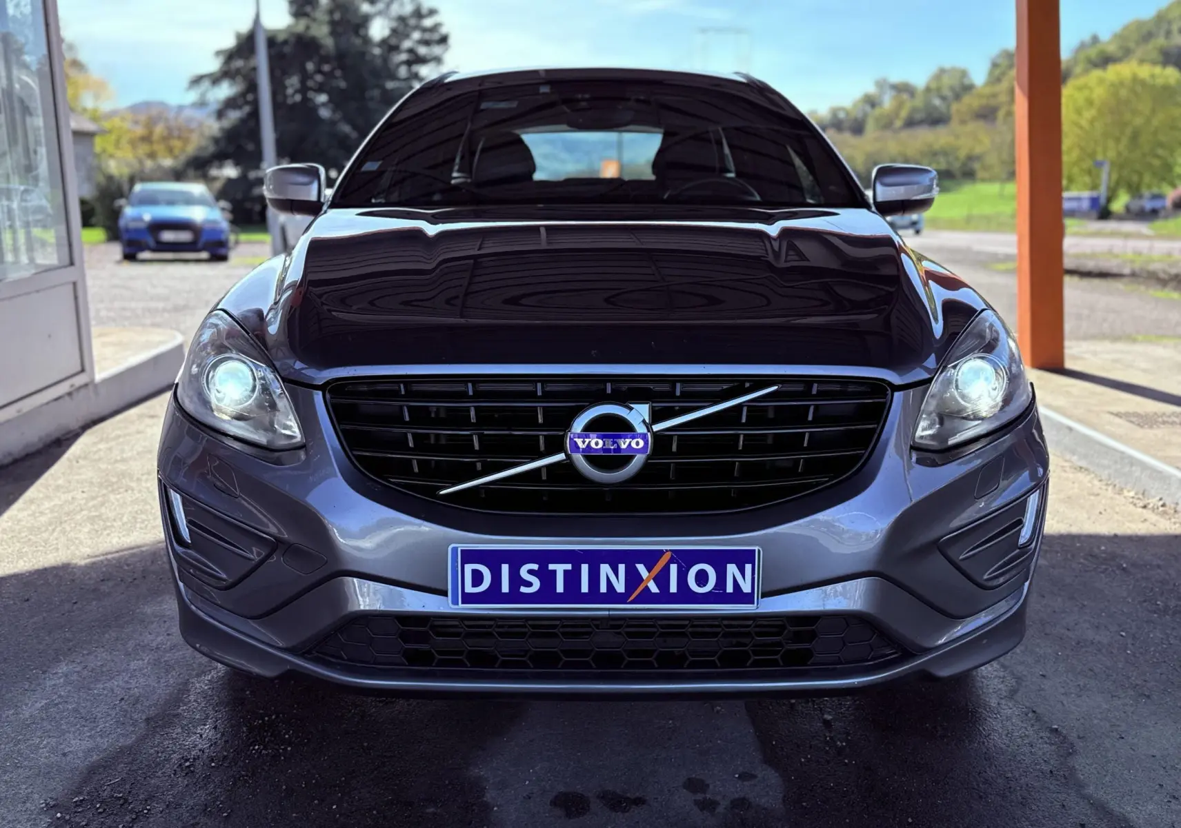 Vue frontale d'un Volvo XC60 gris 2016 avec phares allumés et plaque d'immatriculation Distinxion visible.