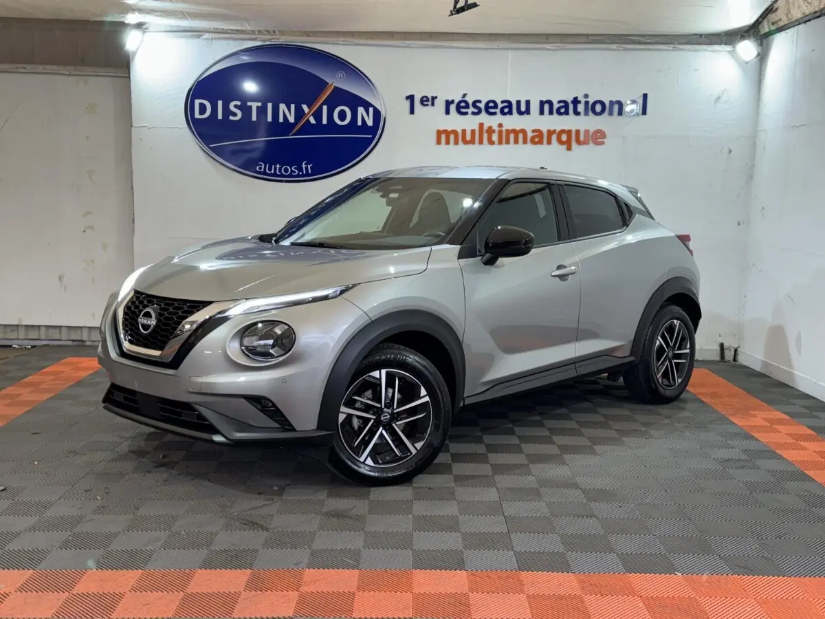 Nissan Juke gris clair vu en 3/4 avant droit, avec jantes alliage noires et calandre V-Motion chromée.
