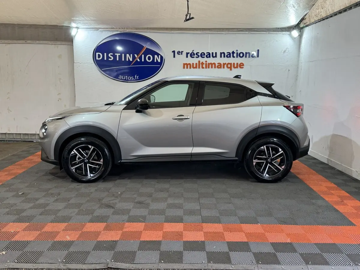 Vue de profil côté gauche d'un Nissan Juke gris clair 2025 avec jantes alliage et toit noir dans un showroom.