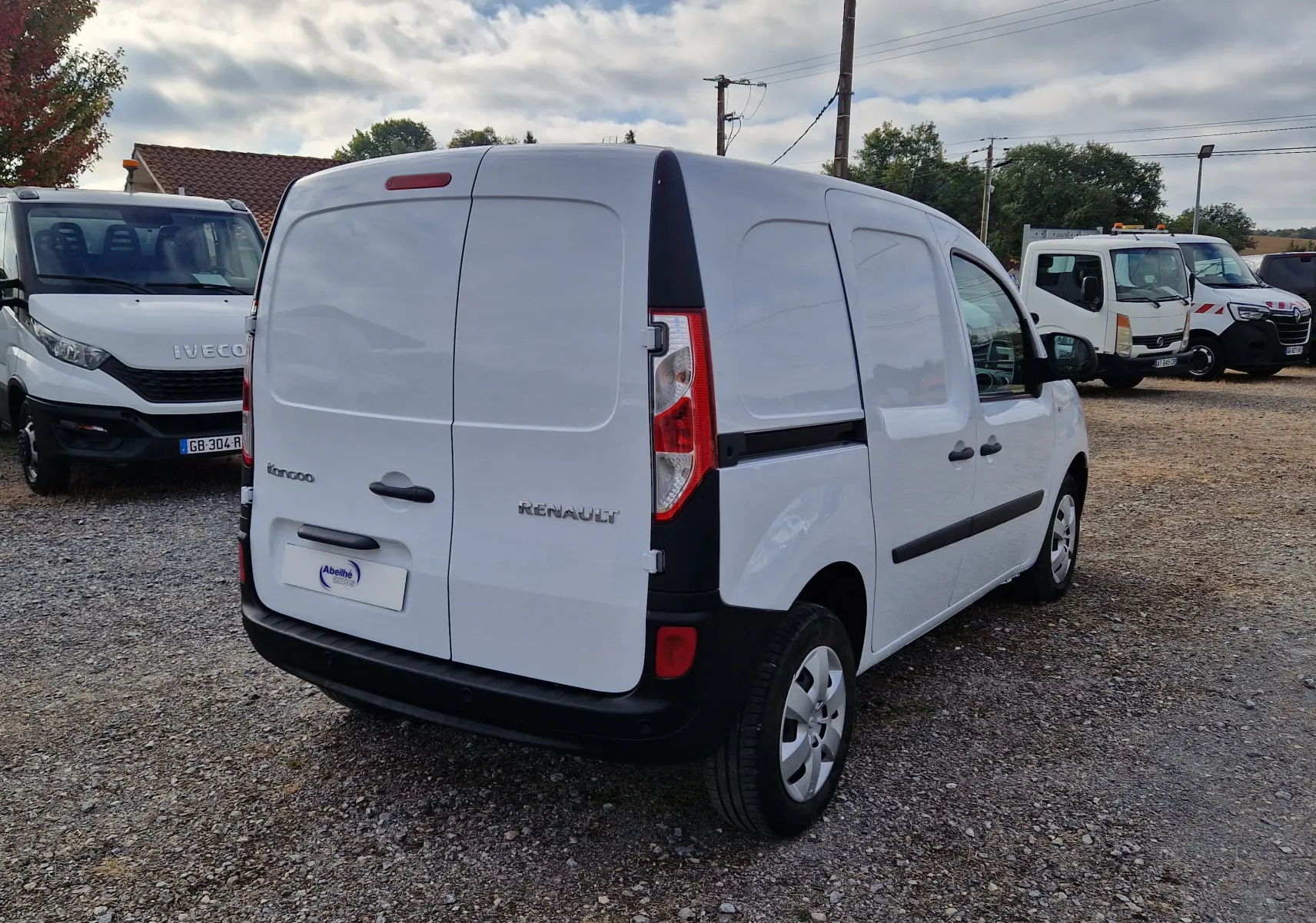 Vue 3/4 arrière droite d’un Renault Kangoo blanc utilitaire avec portes arrière battantes sur un parking gravelé.