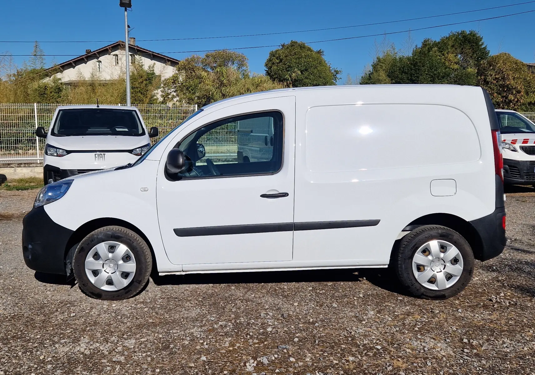 Vue de profil côté gauche d’un Renault Kangoo blanc 2021 utilitaire avec poignées et protections noires sur fond extérieur ensoleillé.