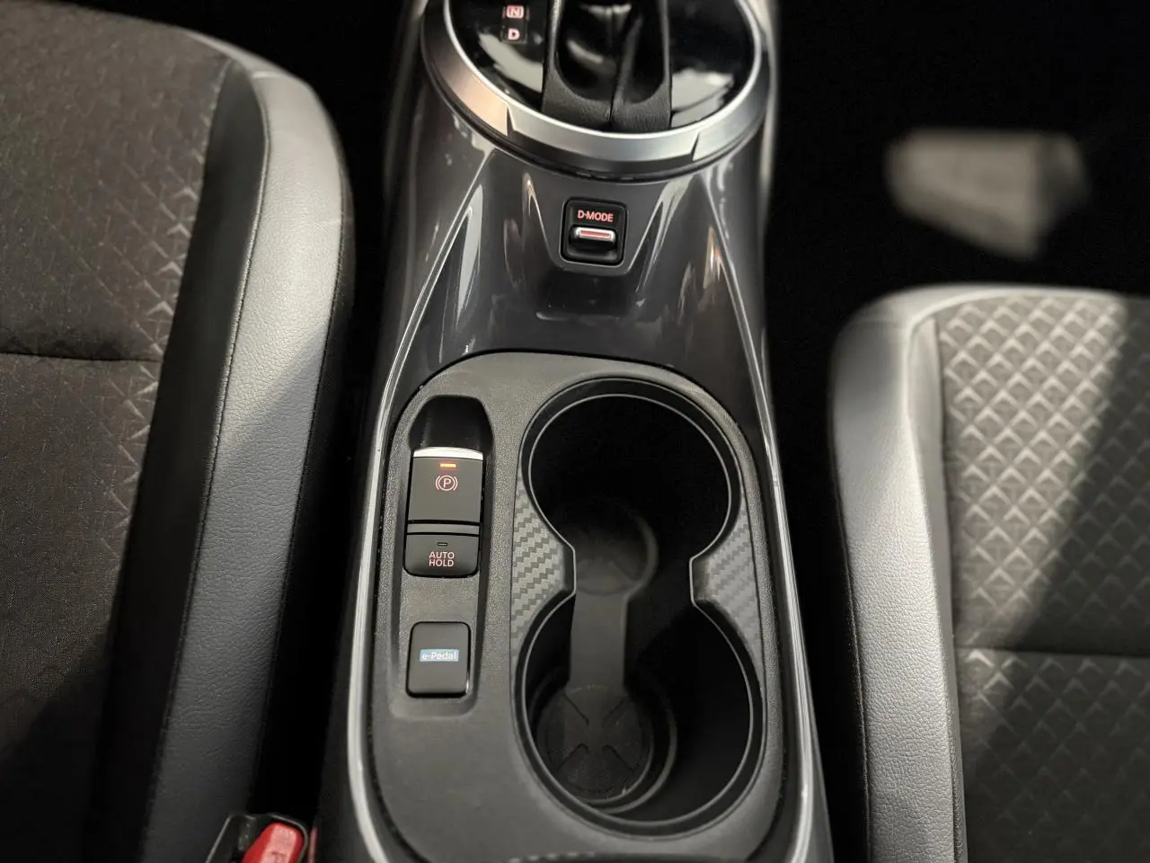 Gros plan sur la console centrale noire du Nissan Juke Hybrid 2024 avec porte-gobelets et commandes e-Pedal et frein électrique.