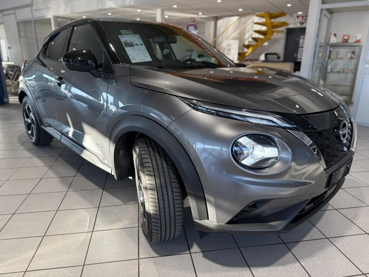 Nissan Juke Hybrid gris métallisé en 3/4 avant droit, avec phares LED et calandre noire en showroom.