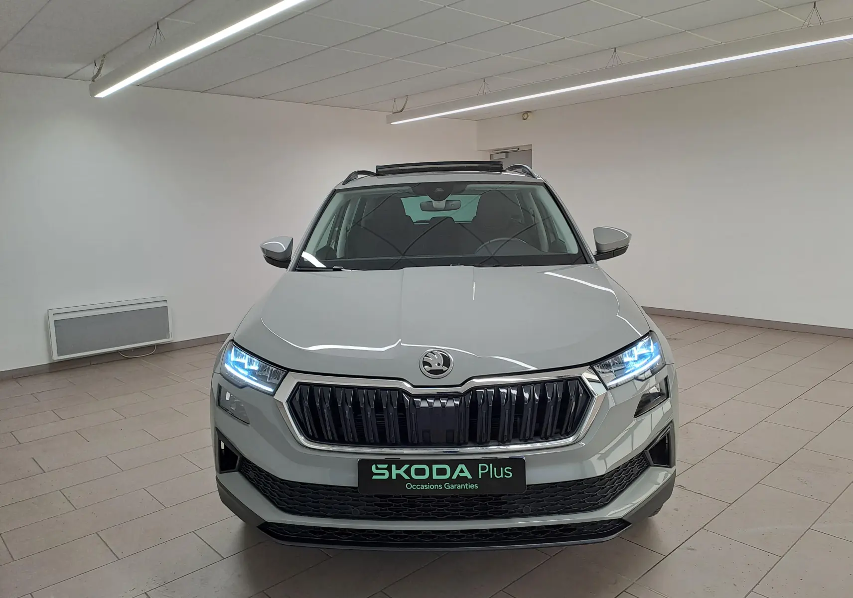 Vue frontale d'un Skoda Karoq gris perle 2023 avec phares allumés et toit ouvrant panoramique visible en intérieur.