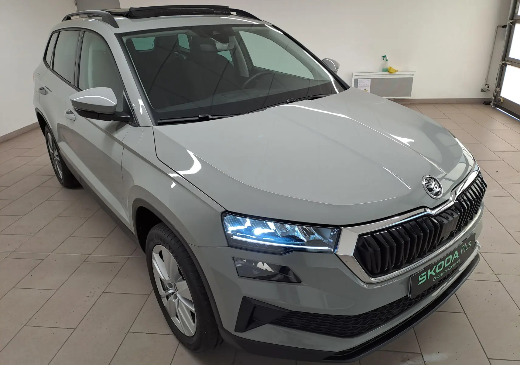 Vue 3/4 avant gauche d'un Skoda Karoq gris perle avec toit ouvrant panoramique et phares allumés en intérieur.