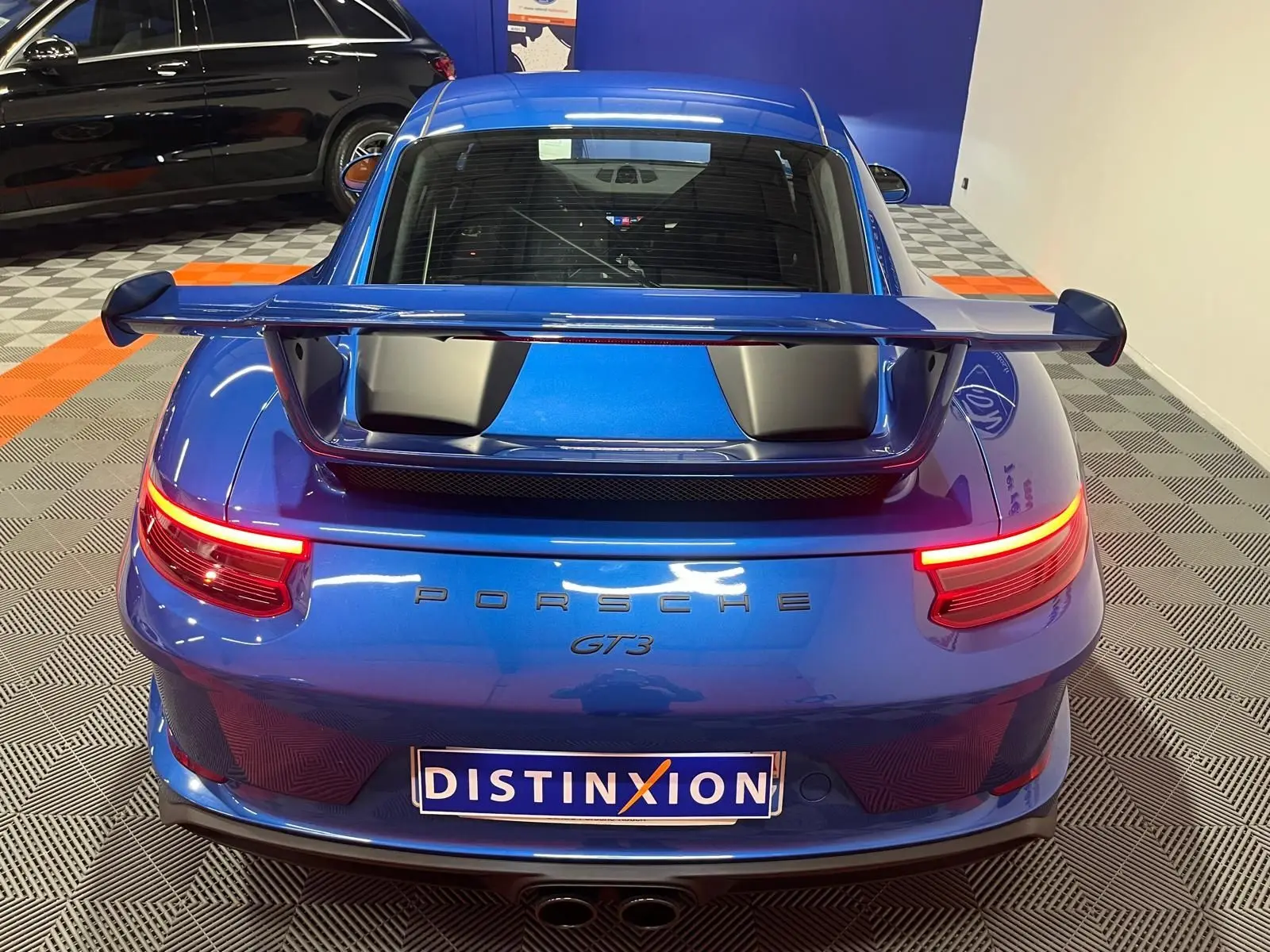 Vue arrière d'une Porsche 911 GT3 bleu saphir avec aileron large et feux arrière allumés en intérieur showroom.
