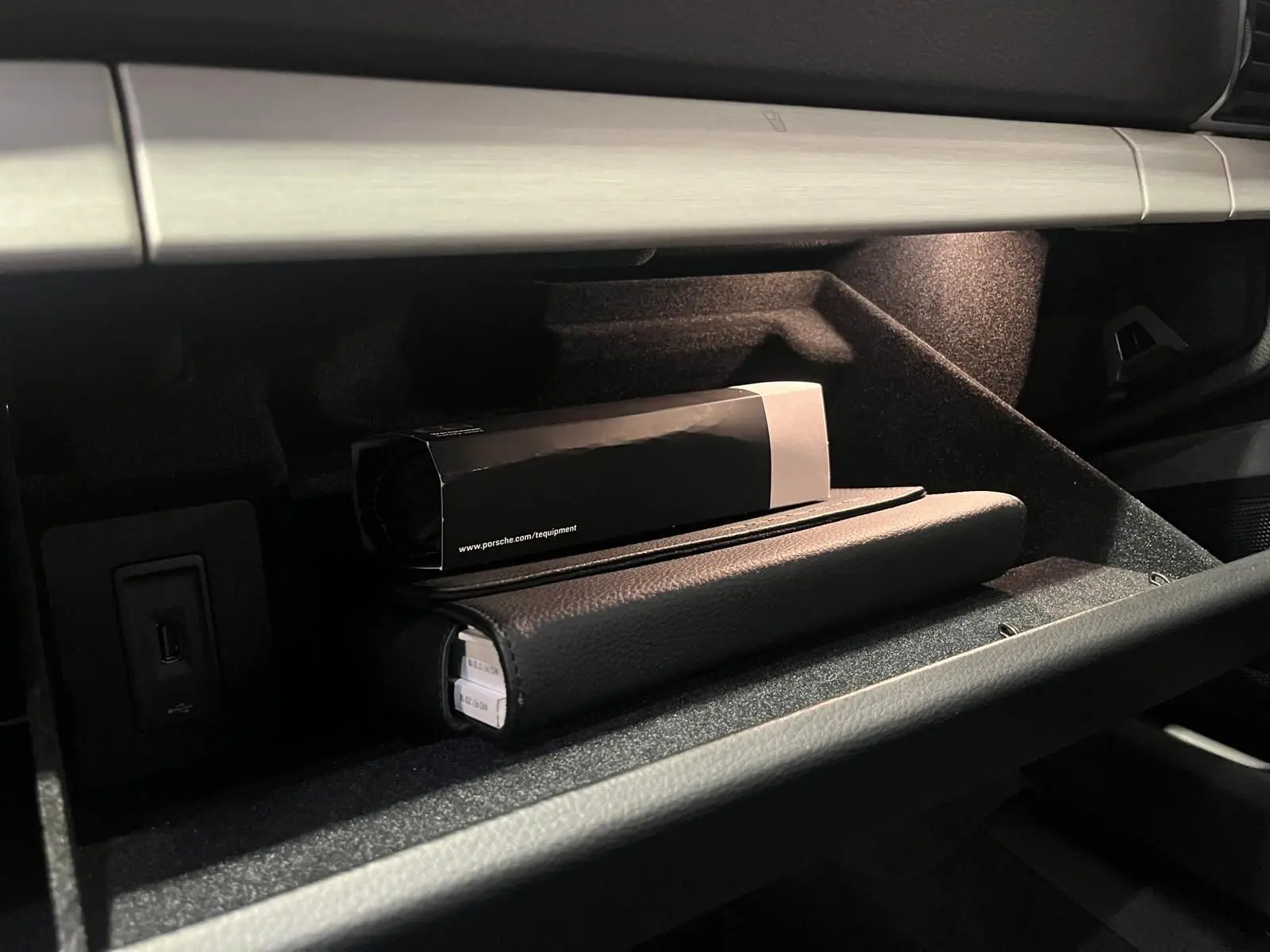 Gant de boîte à gants ouvert avec éclairage intérieur, montrant un manuel Porsche et une boîte noire.