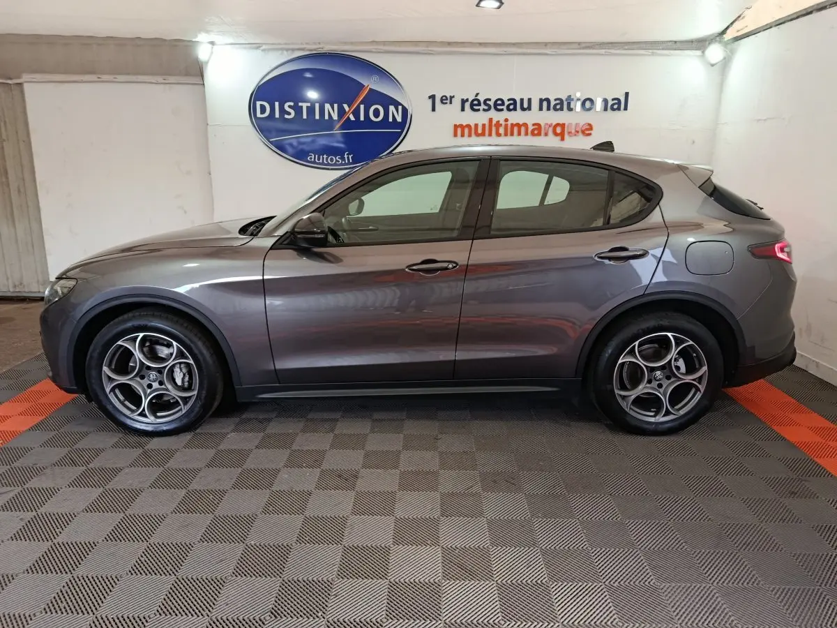 Vue de profil côté gauche d'un Alfa Romeo Stelvio gris foncé, avec jantes distinctives à cinq cercles et feux arrière allumés.