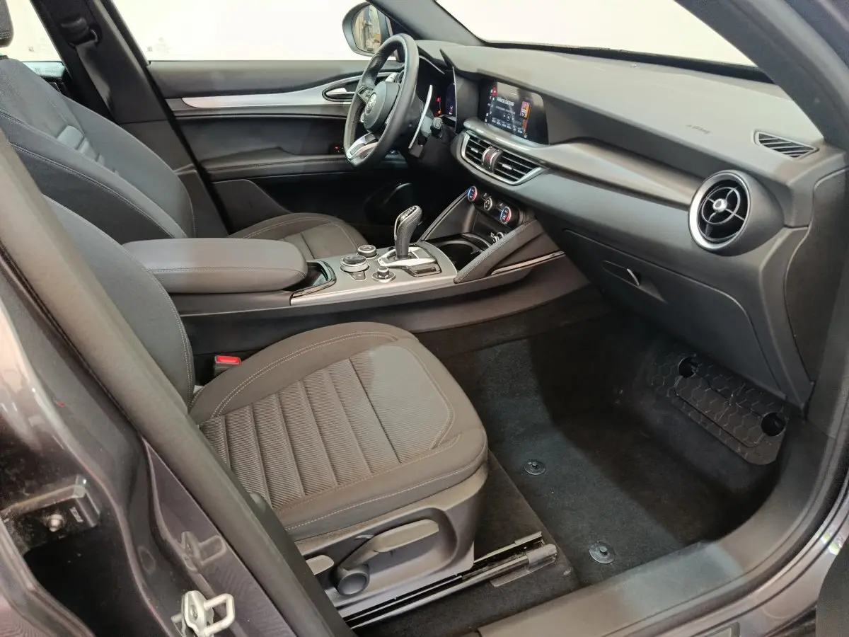 Vue intérieure côté passager de l'Alfa Romeo Stelvio gris foncé 2024, avec tableau de bord moderne et sièges tissu noirs.
