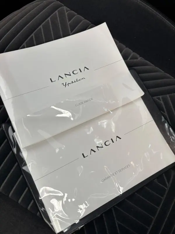 Image montrant deux manuels Lancia Ypsilon posés sur un siège en tissu gris foncé à motifs géométriques, sans vue du véhicule.