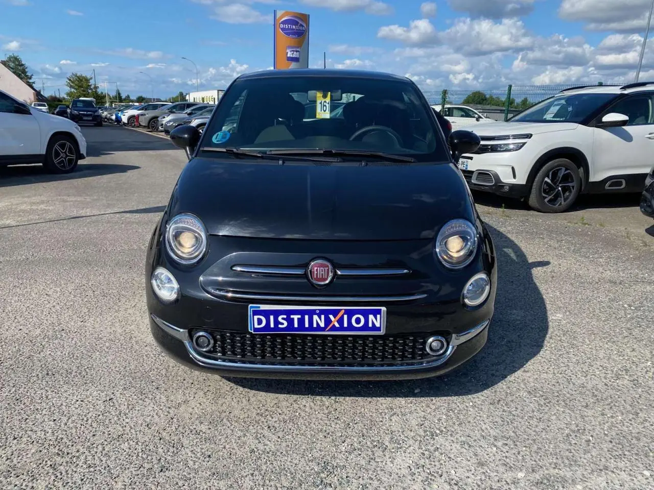 Vue frontale d'une Fiat 500 noire 2024 avec phares allumés et plaque Distinxion sur un parking extérieur.