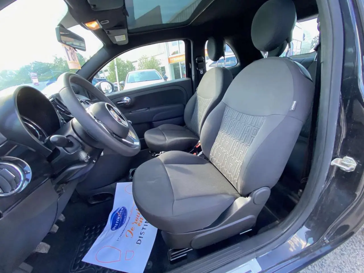 Intérieur côté conducteur d'une FIAT 500 noire 2024, sièges noirs avec motif FIAT, volant et toit panoramique visibles.