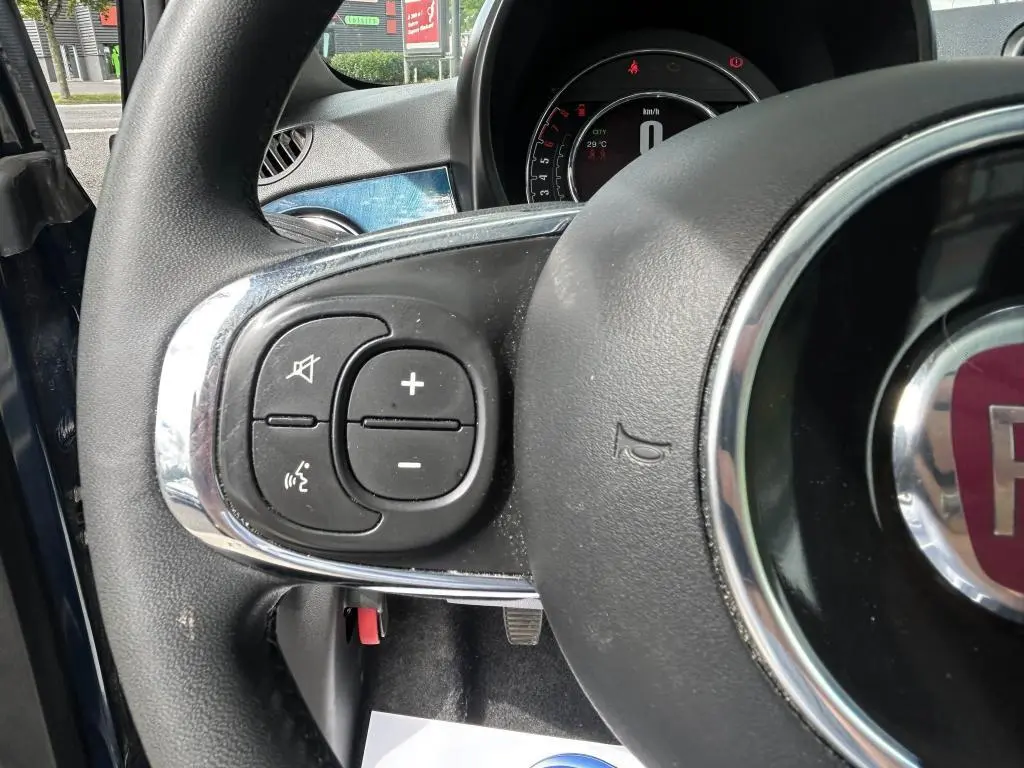 Gros plan sur les commandes au volant gauche du Fiat 500 bleu 2024 avec tableau de bord numérique en arrière-plan.