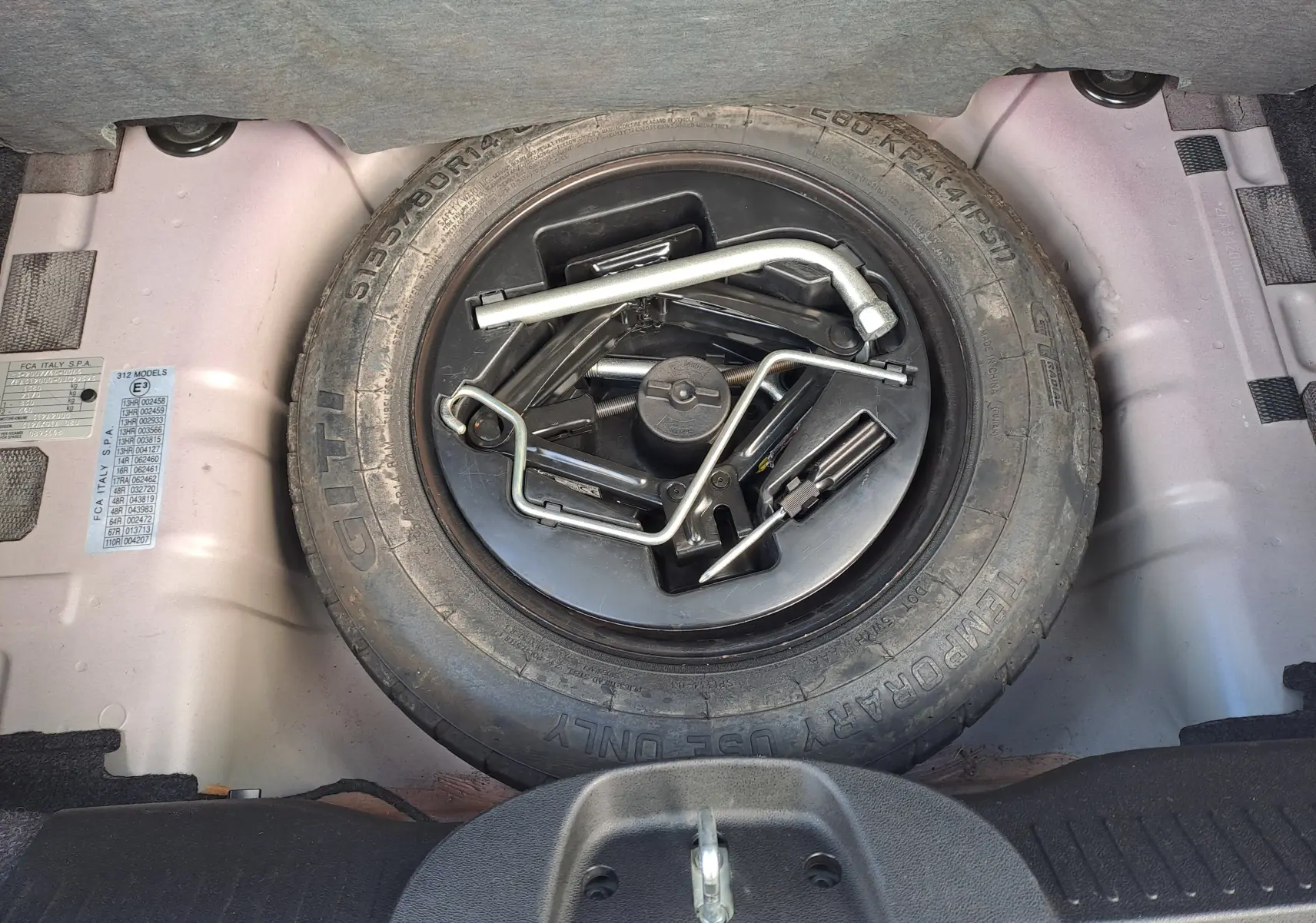 Vue plongeante sur la roue de secours et les outils de bord dans le coffre d'une FIAT 500 Opera Bordeaux Métal 2020.