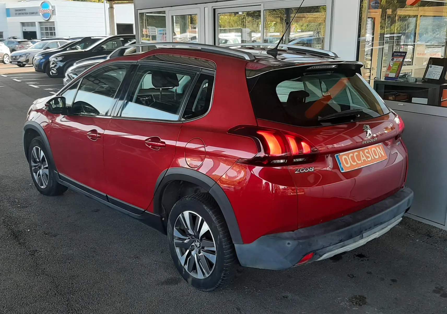 Vue 3/4 arrière droite d'un Peugeot 2008 rouge avec feux arrière allumés et toit noir, stationné devant un concessionnaire.