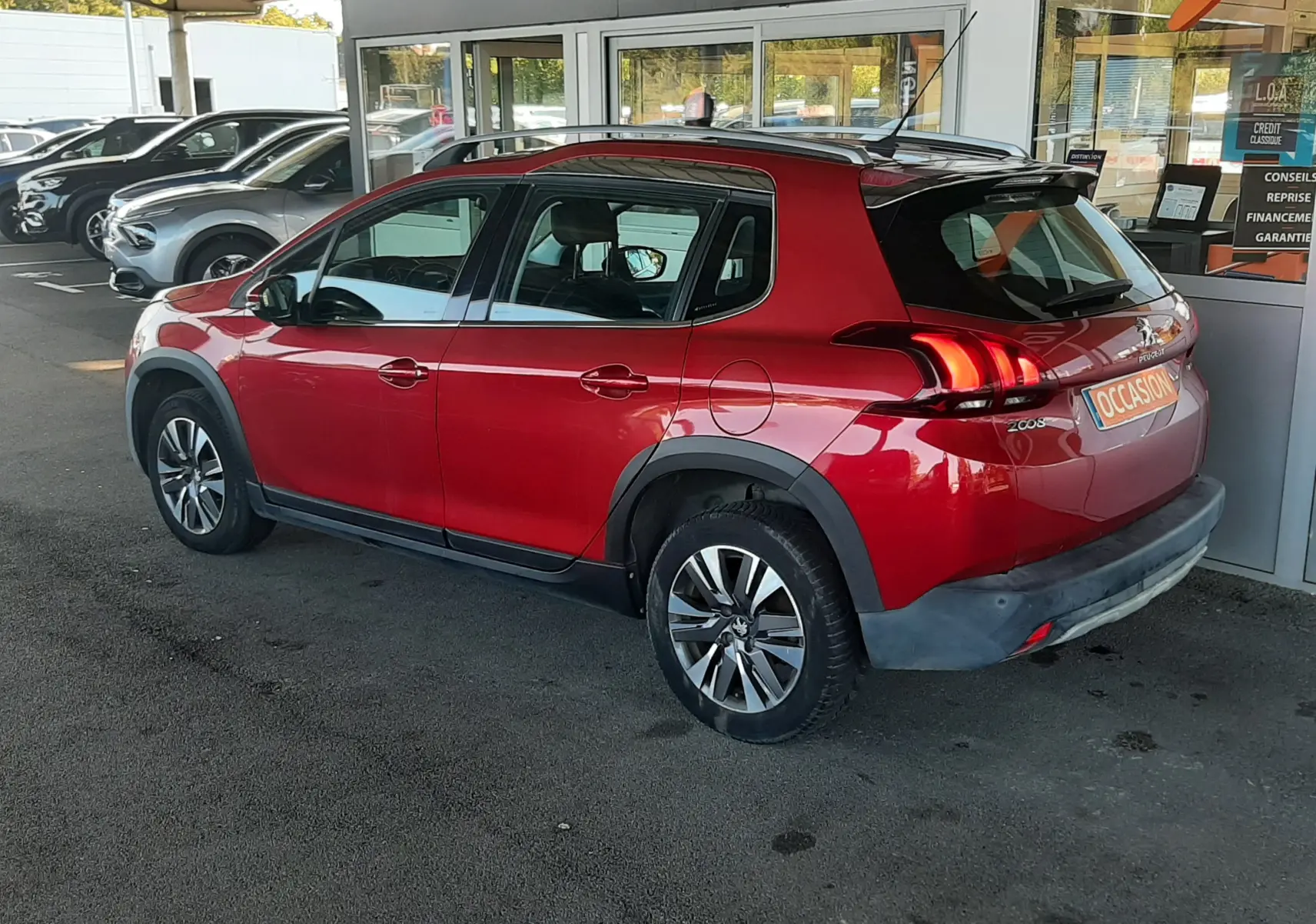 Peugeot 2008 rouge vue de côté droit arrière, stationnée sous abri devant un bâtiment commercial.
