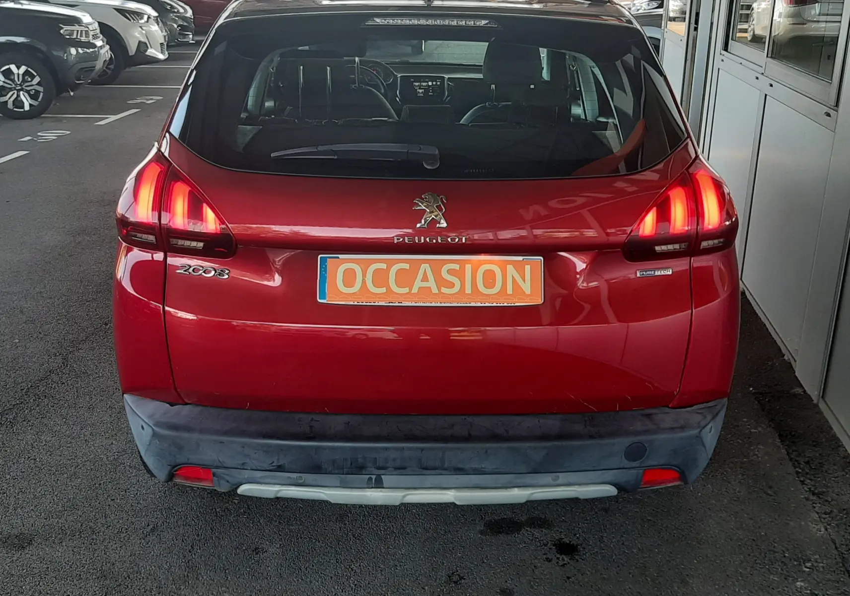 Vue arrière d'un Peugeot 2008 rouge avec feux allumés et plaque "OCCASION" sur un parking extérieur.