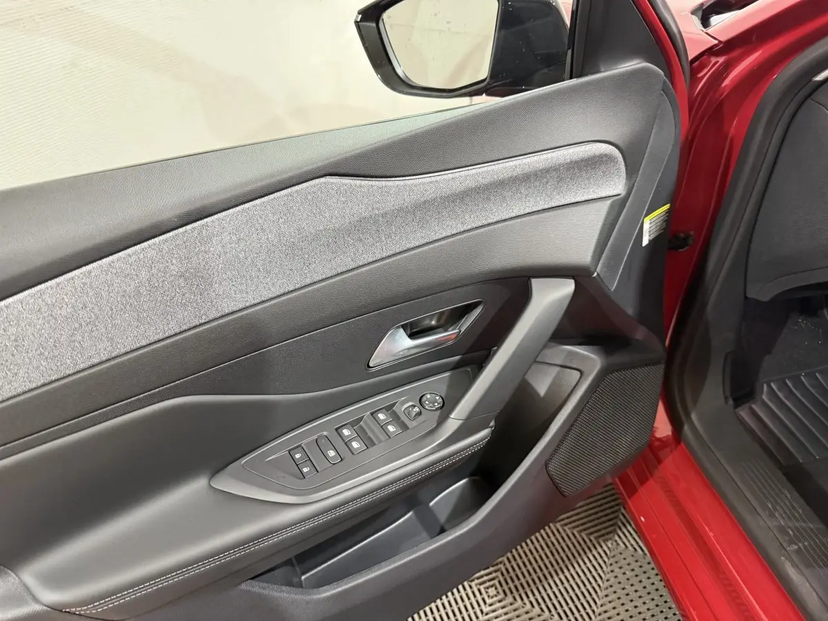 Vue intérieure sur la porte avant gauche rouge d'une Peugeot 308 2025 avec commandes électriques et habillage tissu gris.