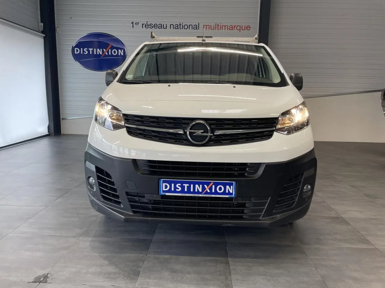 Vue frontale d'un Opel Vivaro blanc avec boucliers noirs et phares allumés dans un showroom.