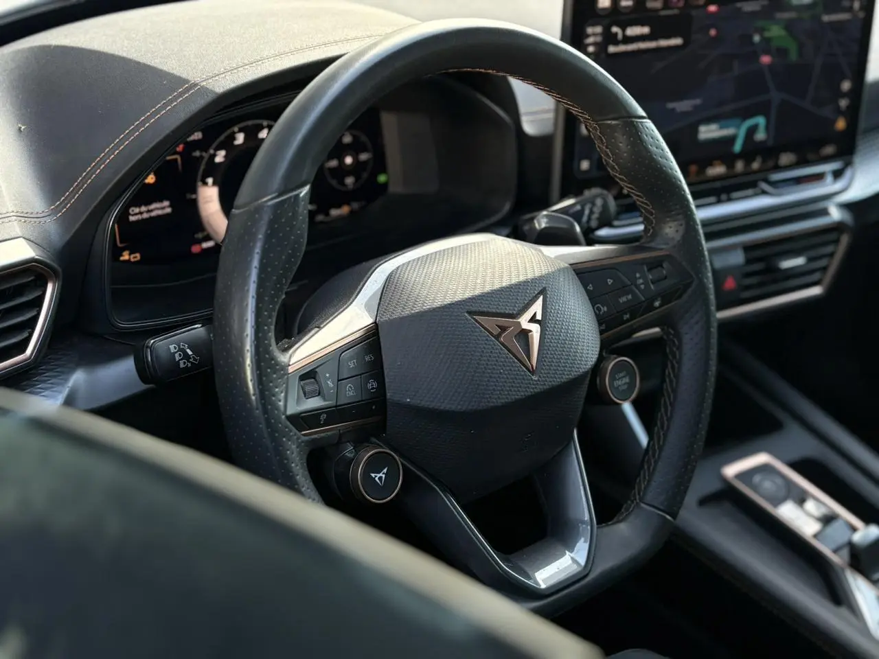 Vue rapprochée du volant cuir perforé noir avec logo CUPRA cuivre et commandes intégrées, tableau de bord numérique en arrière-plan.