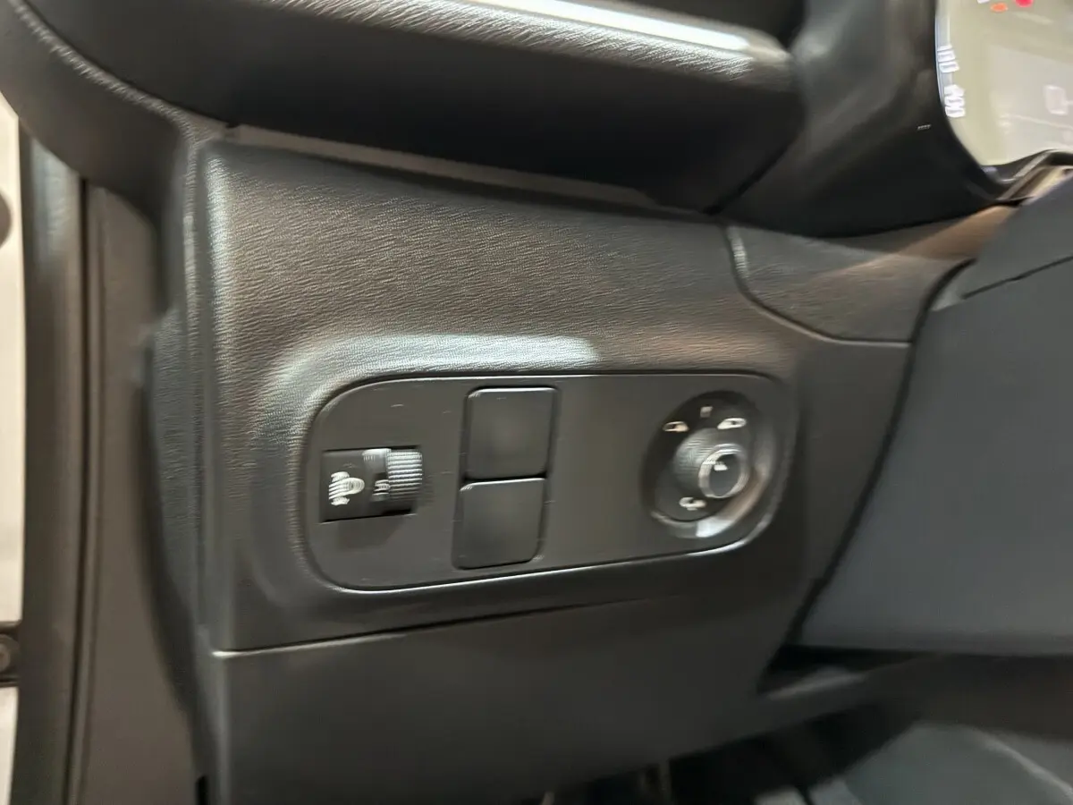 Gros plan sur les commandes de réglage des phares et rétroviseurs côté gauche de la planche de bord noire d'une Citroën C3 blanche.