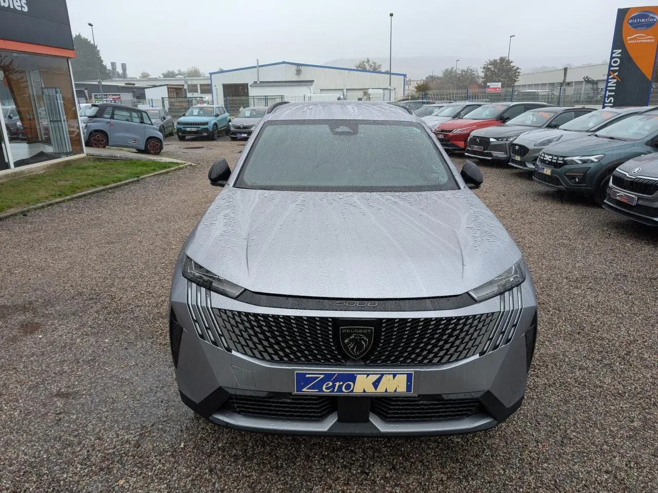 Vue avant d'un Peugeot 5008 gris Artense 2025 avec calandre noire et logo lion central sous la pluie.