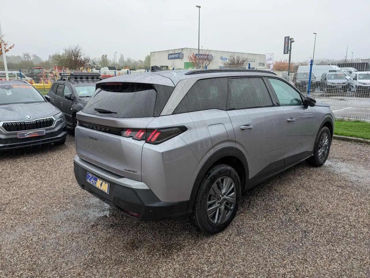 Vue 3/4 arrière droite d'un Peugeot 5008 gris Artense avec feux arrière à LED et jantes alliage 18 pouces sur parking humide.