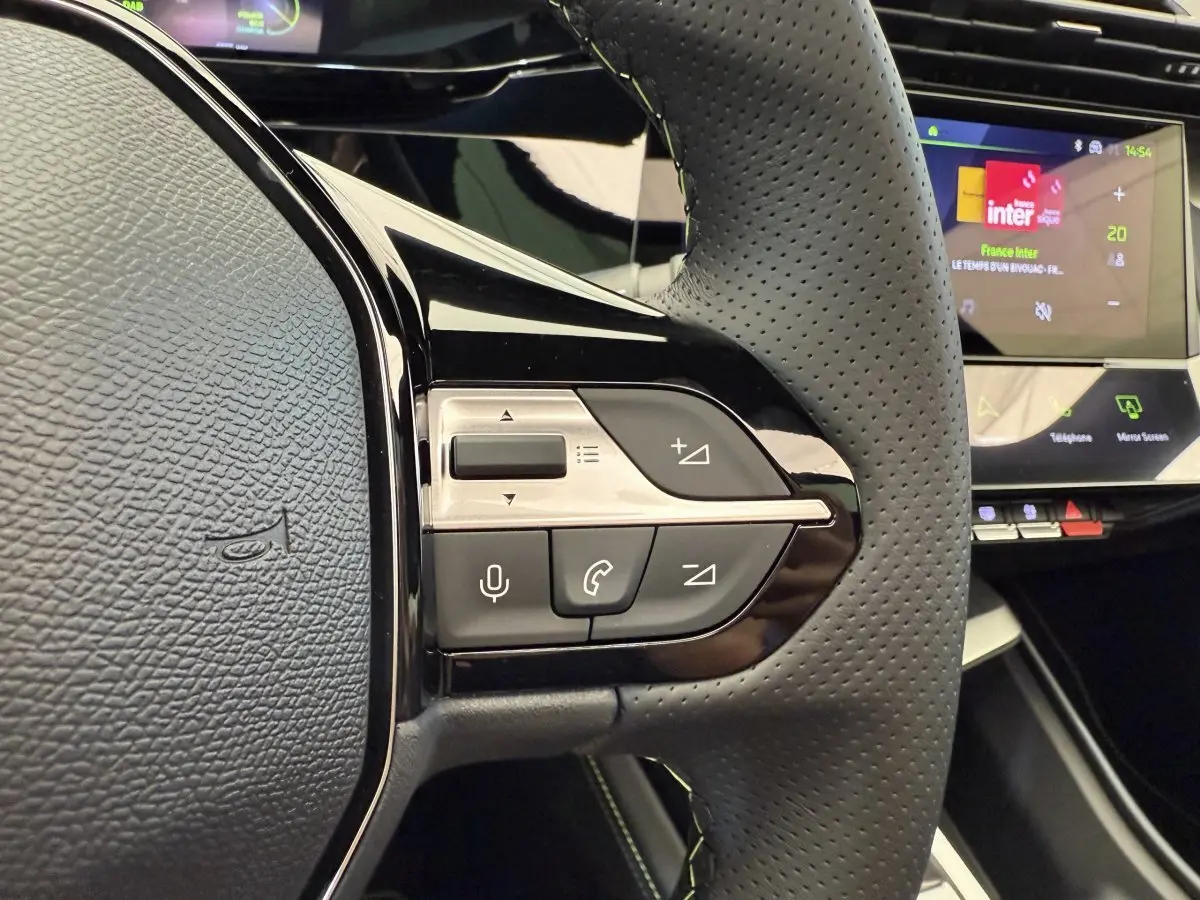 Gros plan sur le volant cuir perforé gris foncé de la Peugeot 308 Hybrid 2024 avec commandes vocales et écran tactile en arrière-plan.