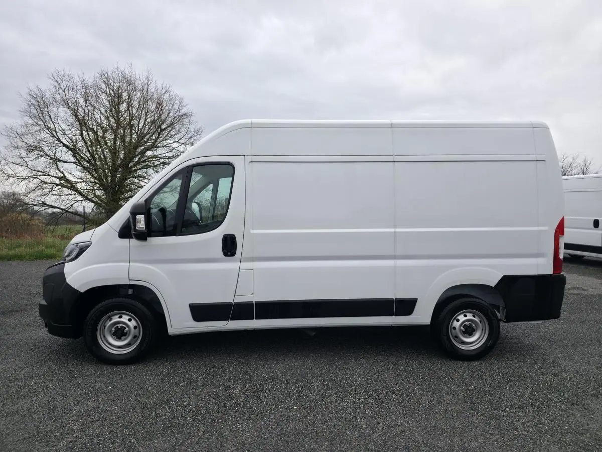 Peugeot Boxer Fourgon blanc en vue de profil côté gauche sur un sol goudronné, fond d'arbre et ciel nuageux.
