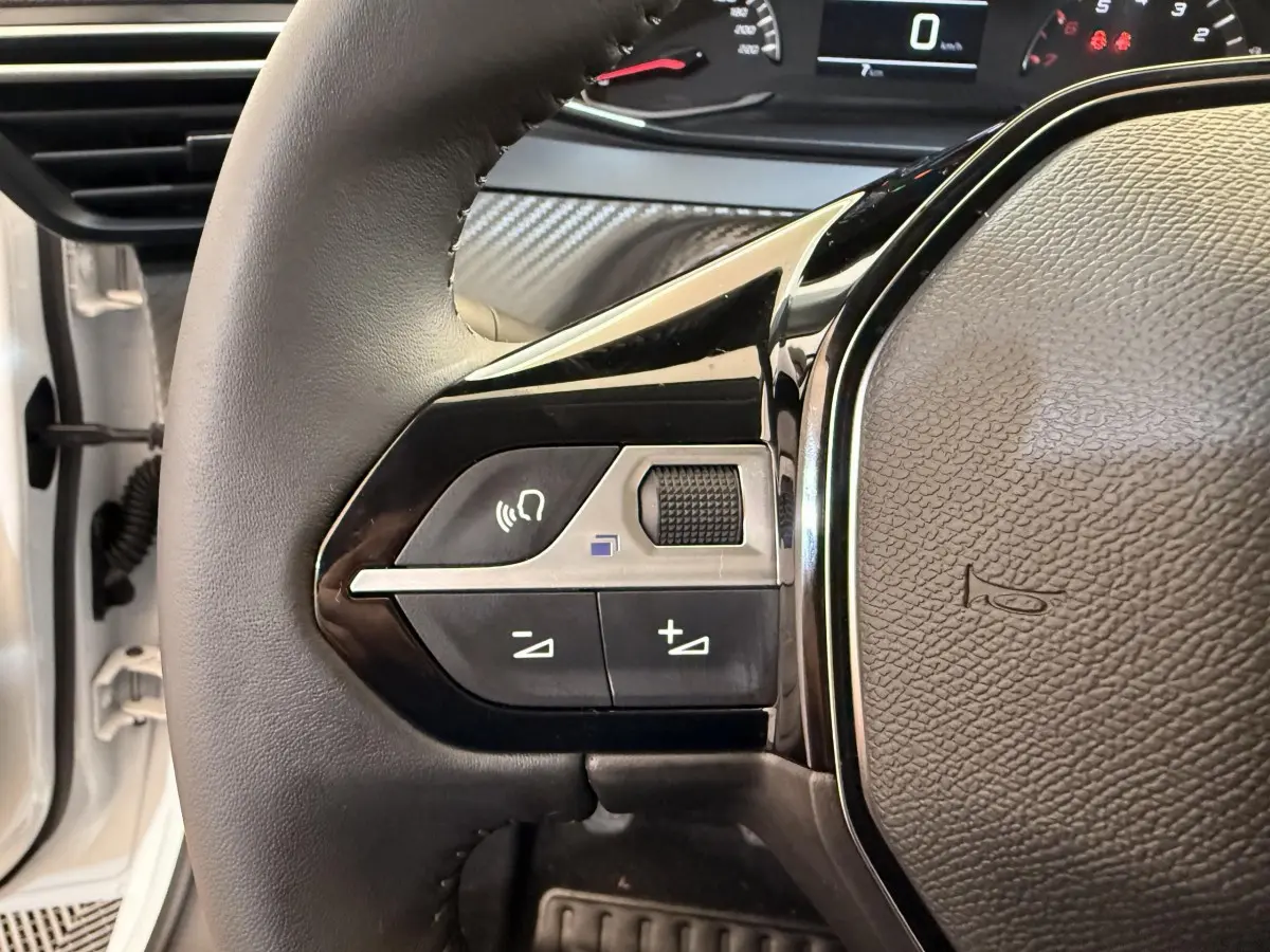 Gros plan sur le volant cuir noir de la Peugeot 208 blanche, avec commandes audio intégrées et tableau de bord partiel visible.