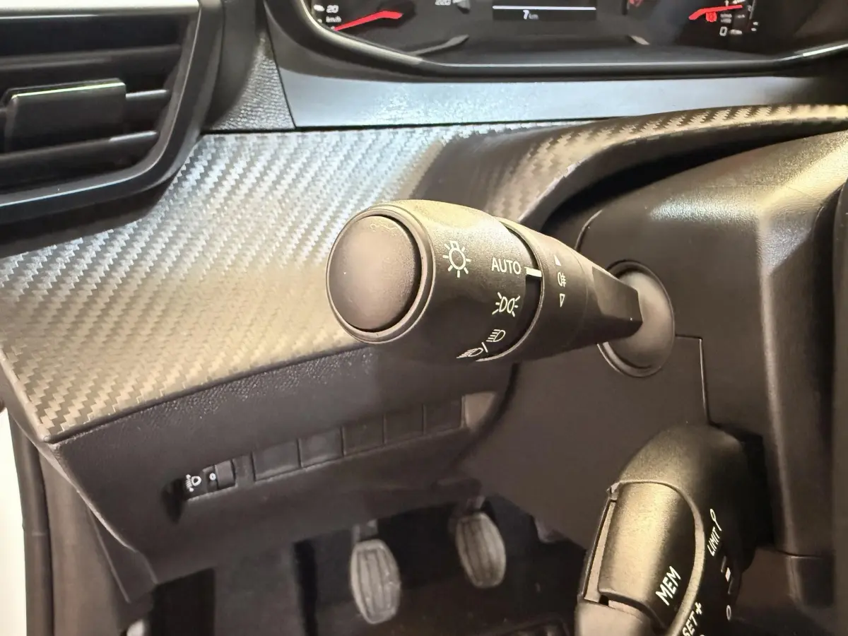Gros plan sur la commande des phares et le volant noir avec insert carbone du tableau de bord de la Peugeot 208 blanche.