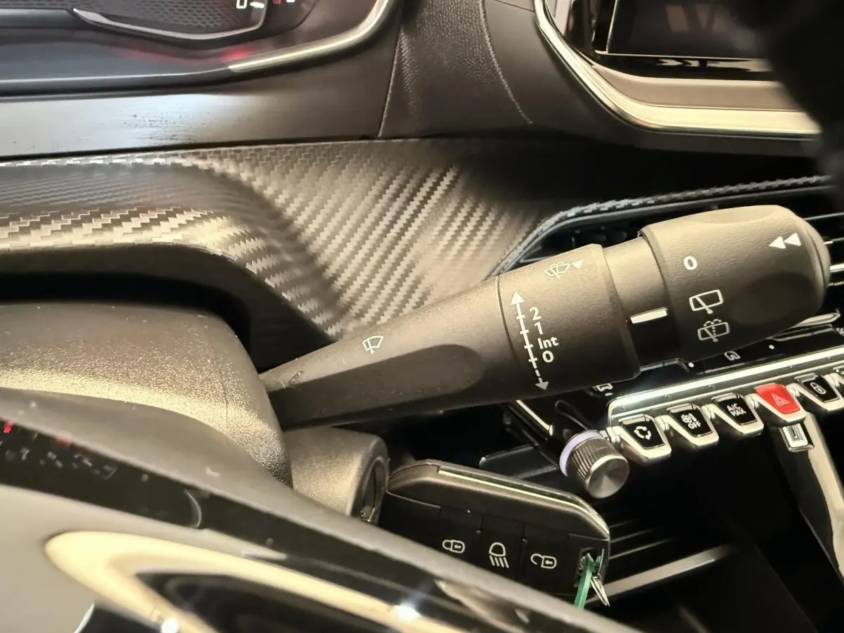 Gros plan sur la commande des essuie-glaces dans l’habitacle noir de la Peugeot 208 blanche, avec tableau de bord à effet carbone.
