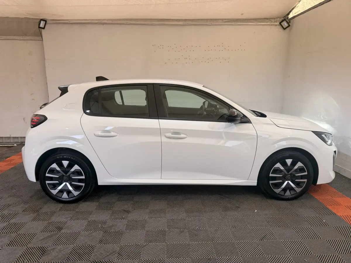Profil droit d'une Peugeot 208 blanche 2025 avec jantes alliage et rétroviseurs noirs dans un garage.