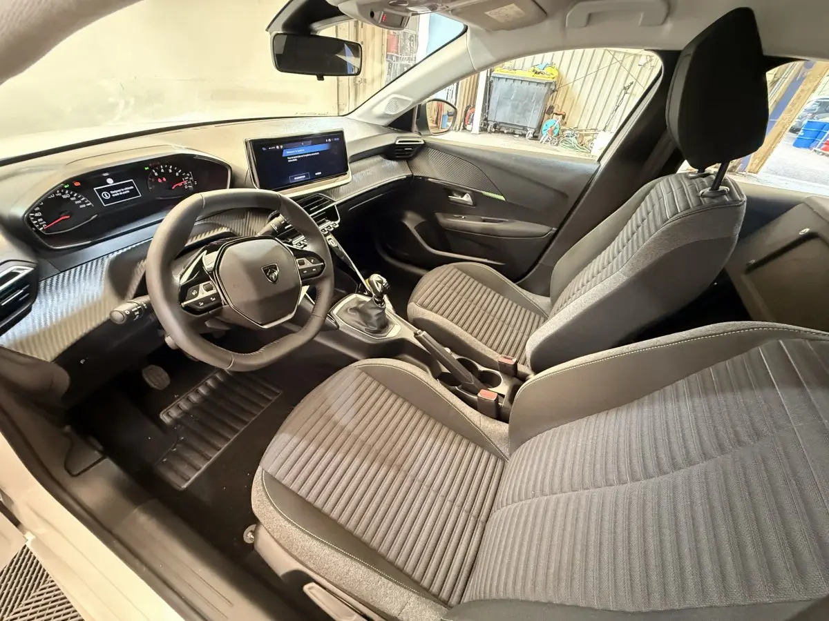 Vue intérieure côté conducteur de la Peugeot 208 blanche, montrant le tableau de bord moderne et les sièges gris à rayures.