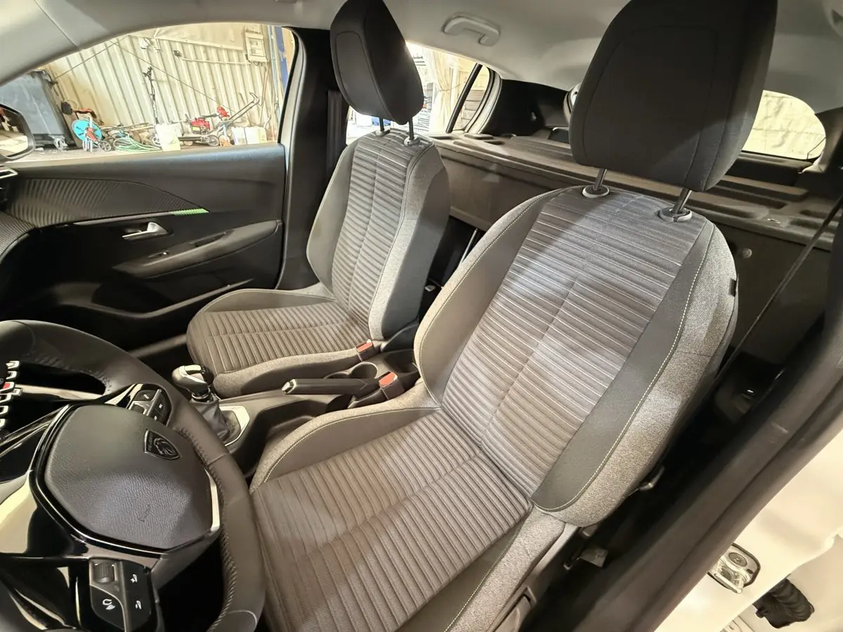Vue intérieure côté conducteur de la Peugeot 208 blanche 2025, montrant les sièges avant en tissu gris et volant cuir.