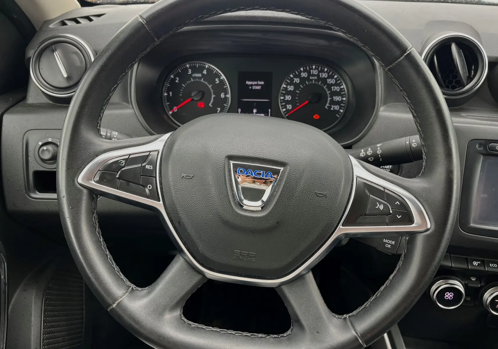 Volant noir de Dacia Duster 1.5 DCI 2018 avec commandes intégrées, tableau de bord et compteurs visibles en arrière-plan.