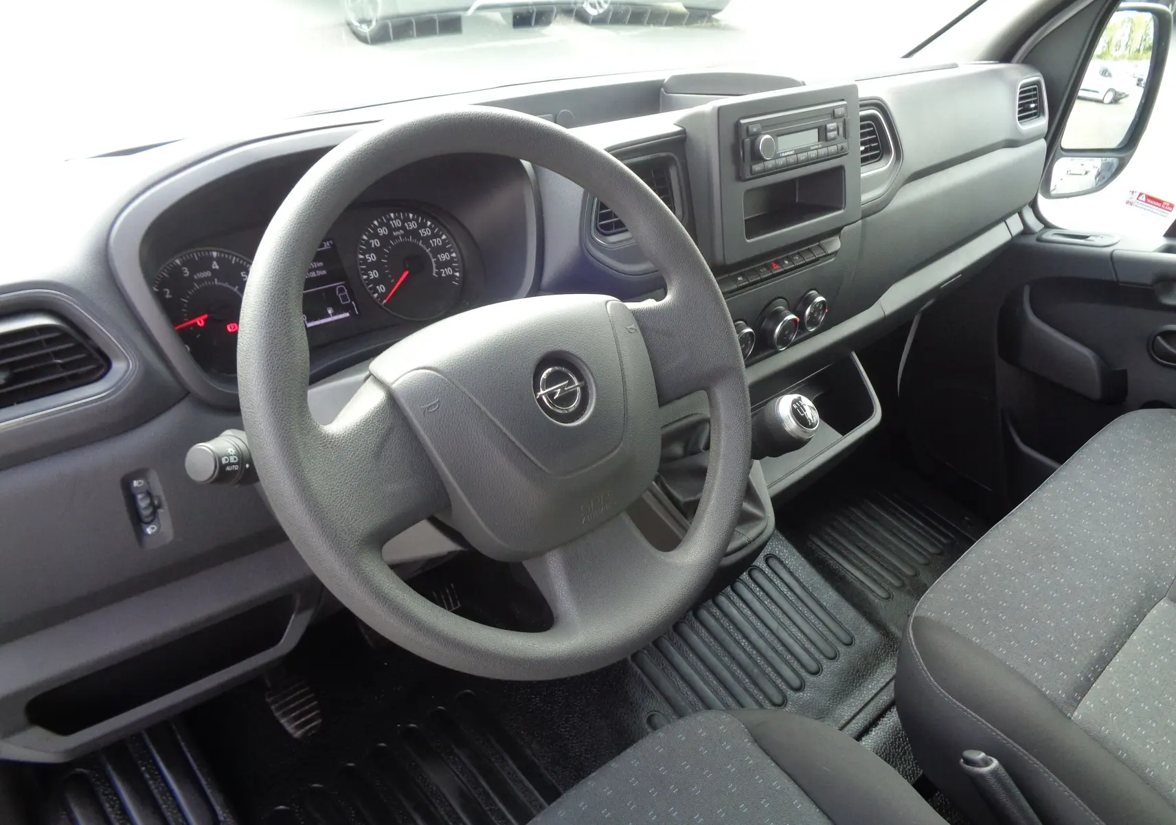 Vue intérieure côté conducteur de l'Opel Movano Fourgon 2022, tableau de bord noir avec volant et levier de vitesses manuelle.