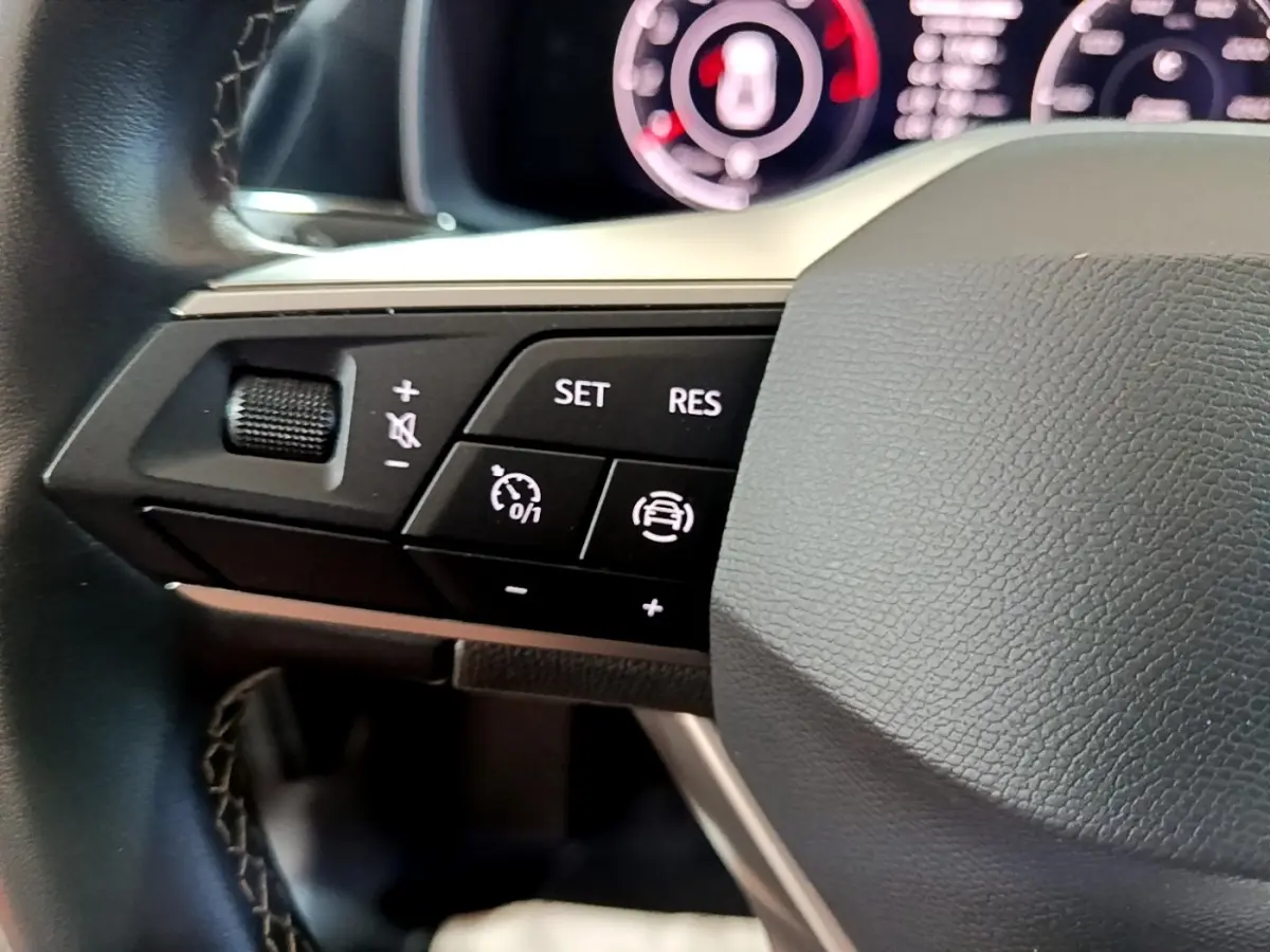 Gros plan sur les commandes à gauche du volant noir d’un SEAT Leon gris clair, avec boutons de régulateur et volume.