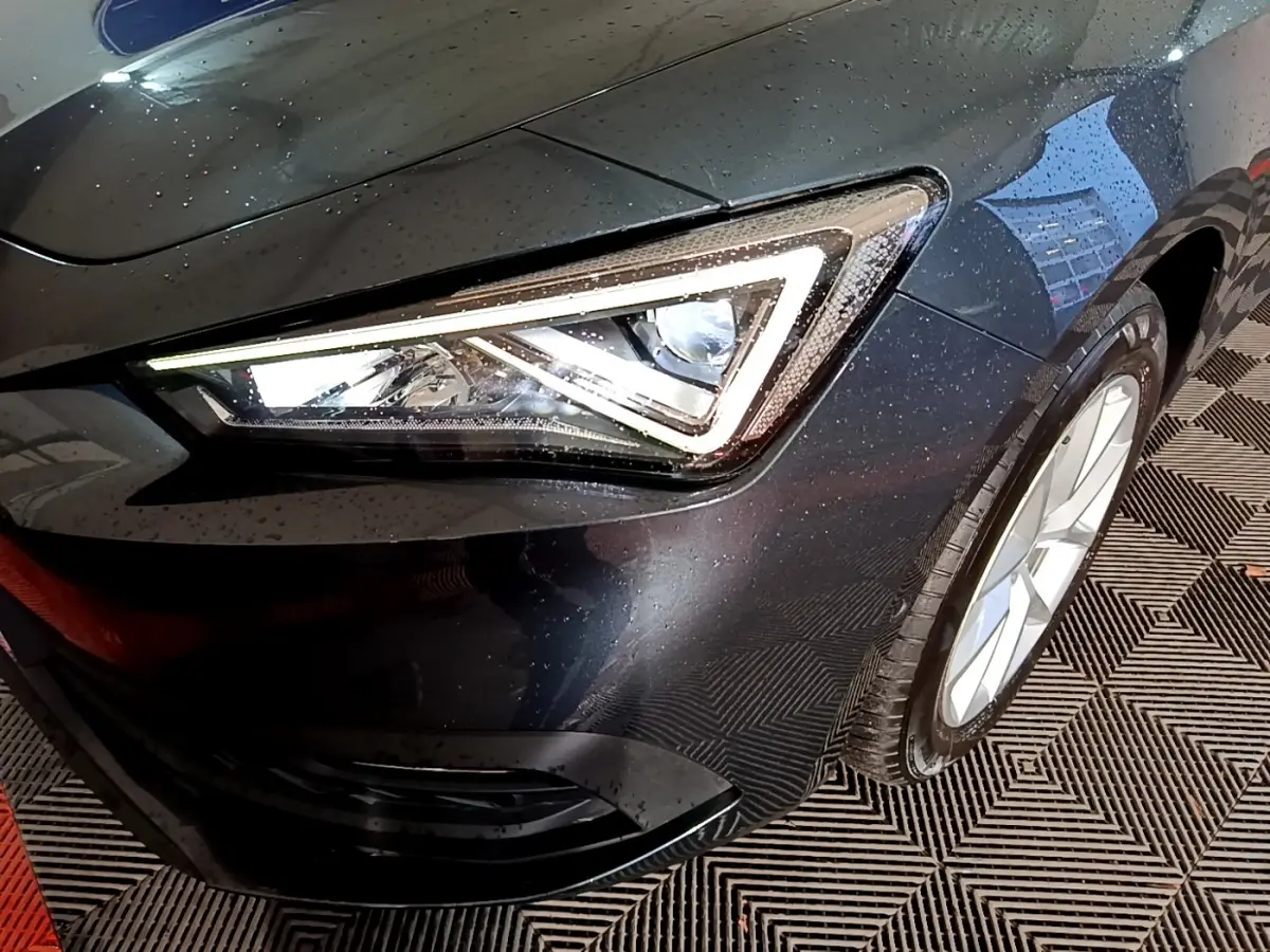 Gros plan sur le phare avant droit allumé d’une SEAT LEON gris clair, avec jante alu visible et carrosserie mouillée.