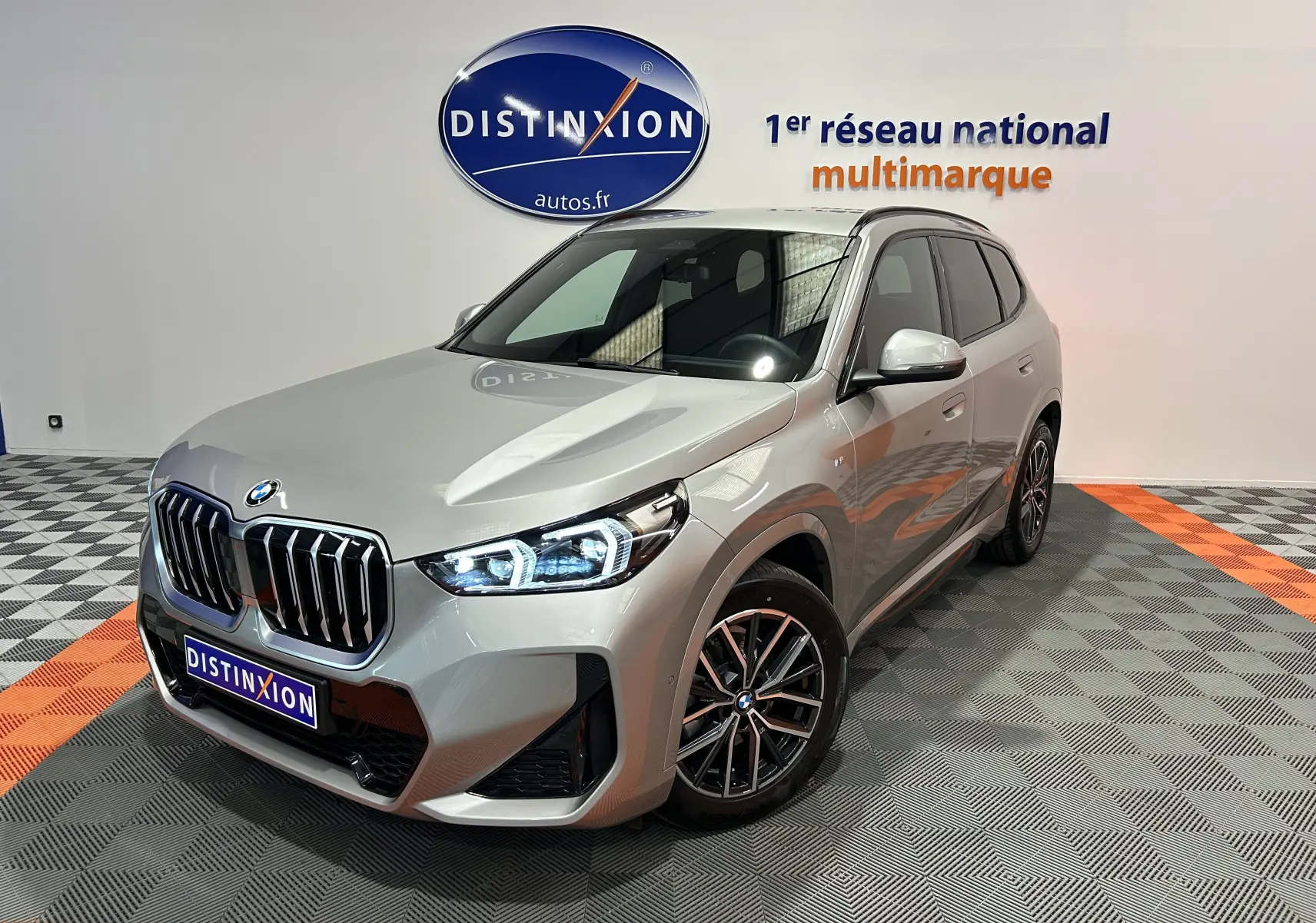 BMW X1 sDrive20d M Sport 2024 en gris Spacesilber, vue 3/4 avant avec phares LED allumés dans un showroom