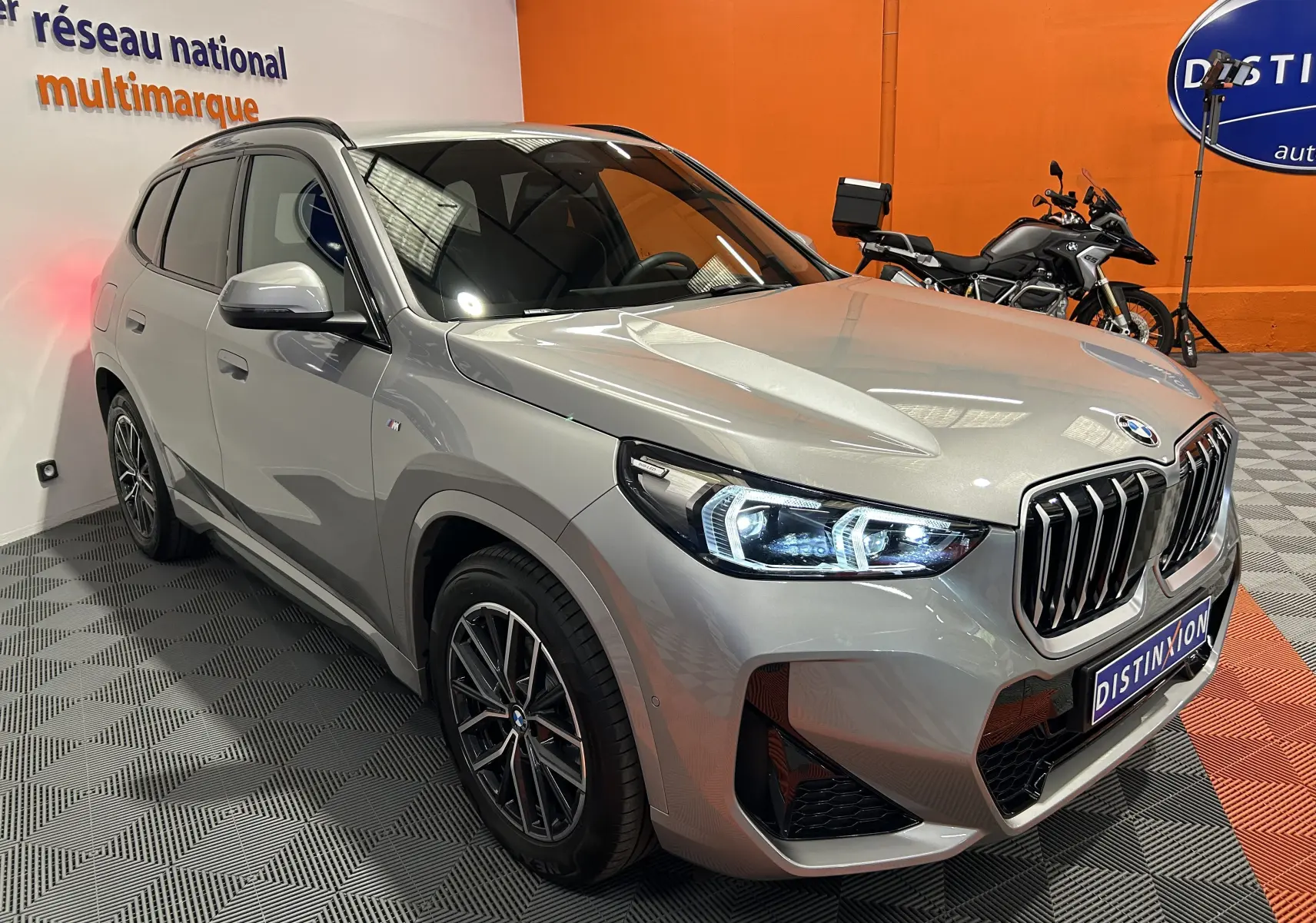 BMW X1 sDrive20d M Sport 2024 en Spacesilber, vue 3/4 avant avec phares LED allumés dans un showroom.