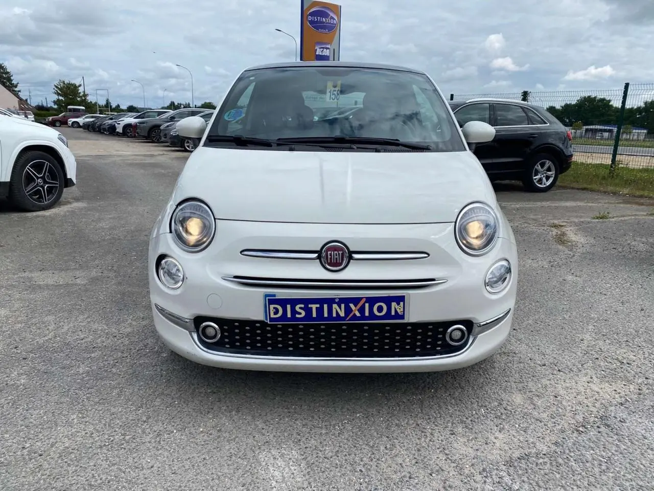 Vue de face d'une Fiat 500 blanche 2024 avec phares allumés et toit panoramique visible en arrière-plan.