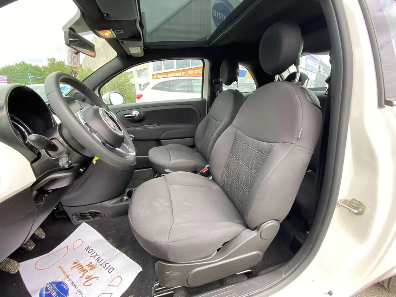 Vue intérieure côté conducteur de la Fiat 500 blanche 2024, mettant en valeur les sièges avant gris et le toit panoramique.