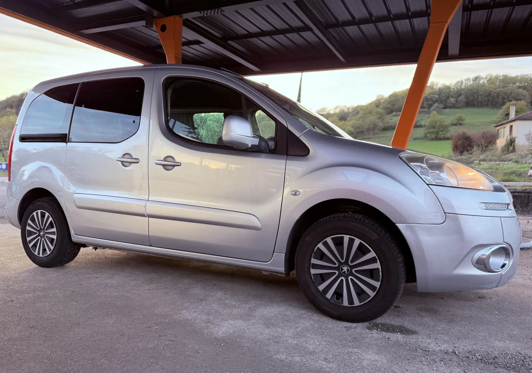 Peugeot Partner Tepee gris vu de profil côté droit, sous abri avec jantes spécifiques et vitres teintées.