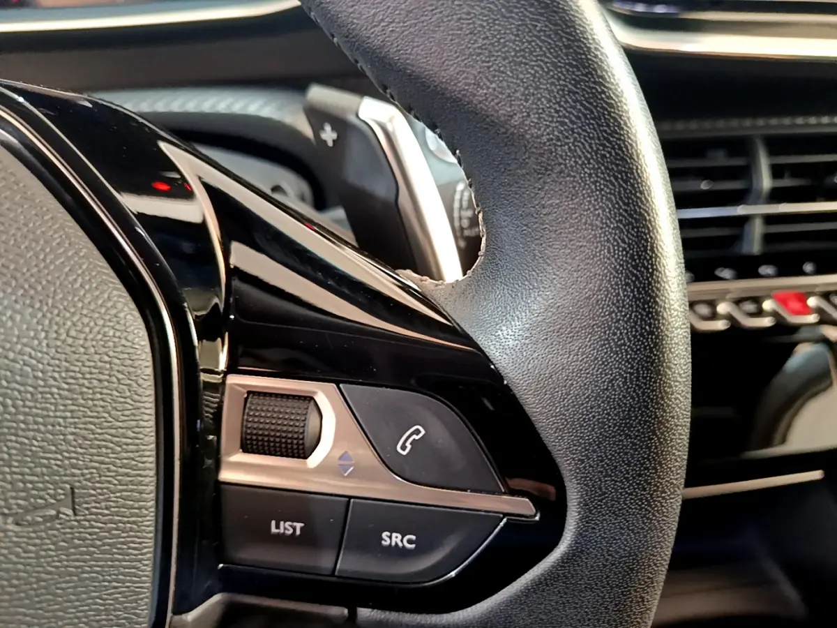 Gros plan sur le volant cuir noir du Peugeot 2008 gris clair, avec commandes audio et tableau de bord en arrière-plan.