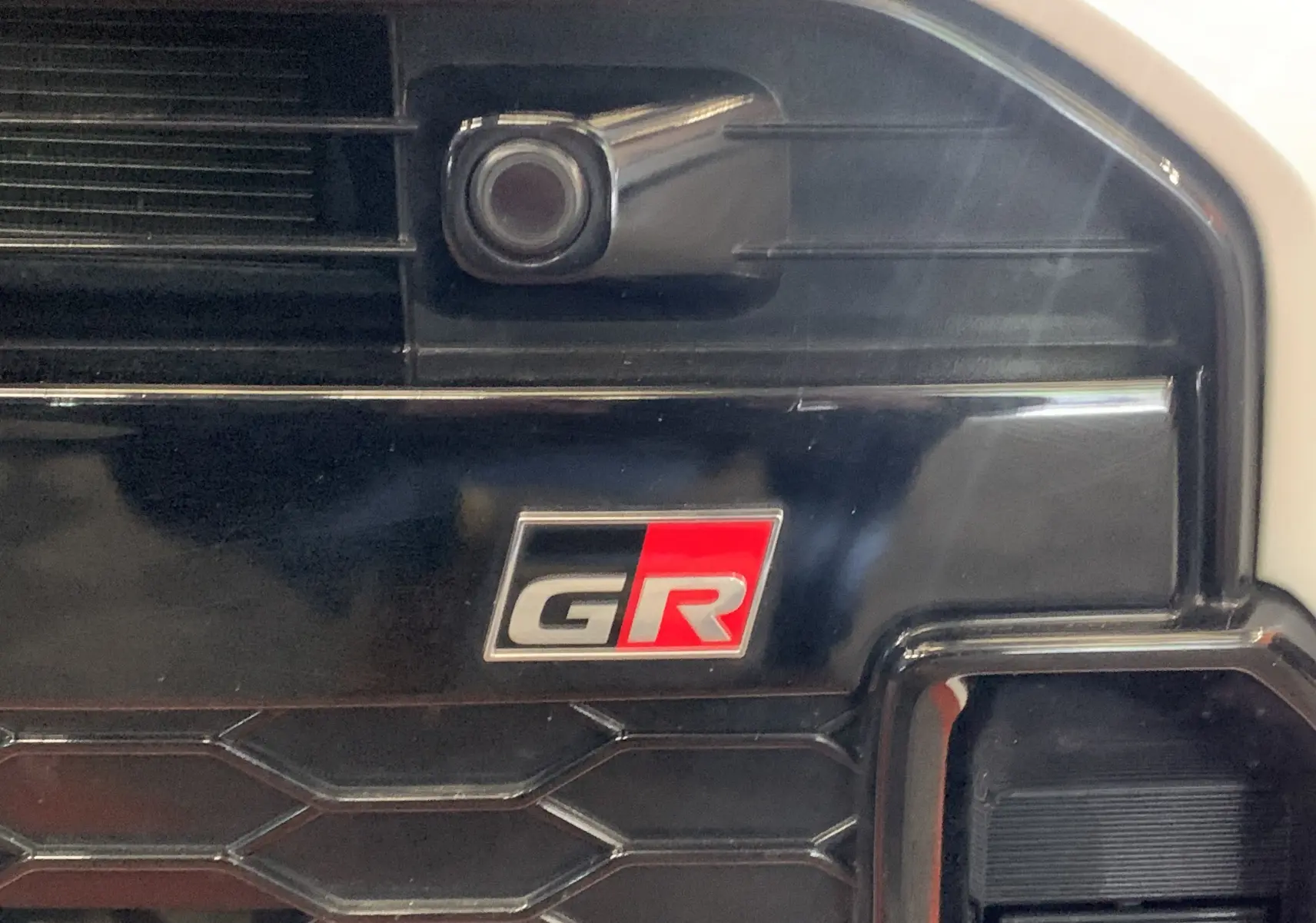 Gros plan sur la calandre noire de la Toyota Yaris 1.6 VVT-I 261 Premium GR avec logo GR rouge et gris.