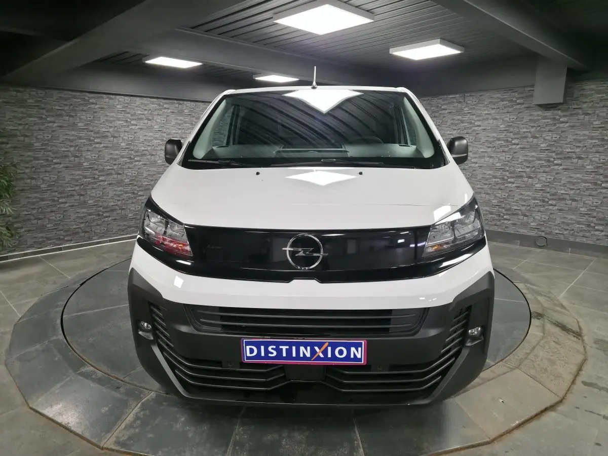Vue avant d’un Opel Vivaro blanc 2025 avec calandre noire brillante et projecteurs intégrés, en intérieur showroom.