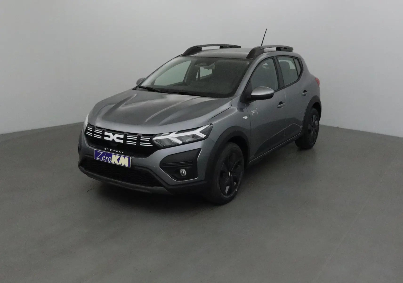 Vue 3/4 avant d'une Dacia Sandero Stepway 2025 gris schiste avec barres de toit et jantes noires.